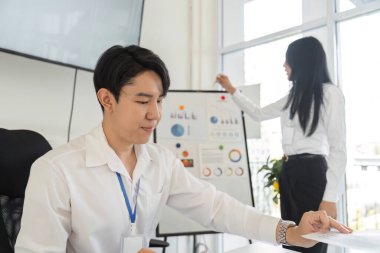 Genç bir profesyonel finansal grafiklere odaklanırken, bir meslektaşı da bu grafiklerin arkasında bir sunum hazırlıyor..