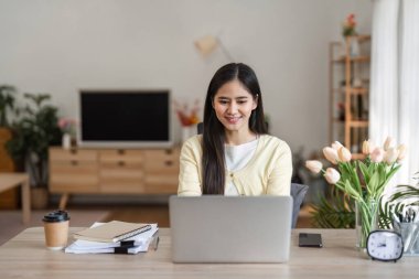 Genç bir kadın bilgisayarında uzaktan çalışırken gülümsüyor rahat bir ofiste, bitkiler ve kişisel eşyalar arasında verimli bir günün tadını çıkarıyor..