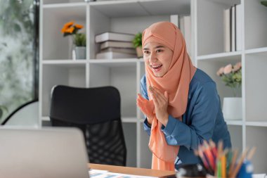 Türbanlı Müslüman bir kadın modern bir ofiste başarılı bir başarıyı sevinçle kutlarken masasında dizüstü bilgisayarla oturuyor..