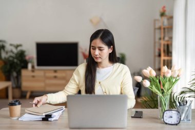 Bir kadın laptopuna odaklanarak notlarını gözden geçirir ve evde etkili bir iş yönetimi sergilediğini gösterir..