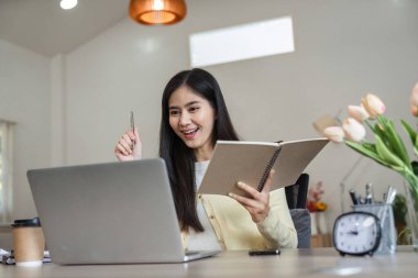 Neşeli bir Asyalı kadın dizüstü bilgisayarına gülümserken bir defter ve kalem tutuyor, üretken ev ofis ortamında coşku ve bağlılık gösteriyor.