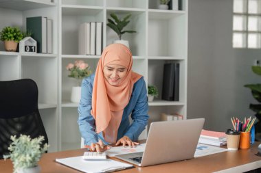 Başörtüsü takan Müslüman bir kadın masasındaki verileri analiz etmek için hesap makinesi kullanır. Çağdaş bir ofis ortamında çalışmalar yapar..