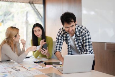 Bir dizüstü bilgisayar kullanan bir üyeyle aktif olarak bir tasarım projesi üzerinde çalışan çeşitli bir ekip, diğerleri ise modern bir ofiste fikirleri paylaşıyor ve inceleme materyallerini paylaşıyor..