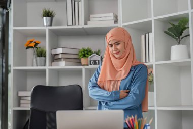 Türbanlı kadın muhasebeci düşünceli bir şekilde duruyor, modern ofis ortamındaki mali raporları yansıtıyor..