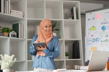 Çeşitli kadın muhasebeci tesettürlü bir kadın, modern ofis ortamında finansal çizelgelerle dolu bir sunum panosunun yanındaki tablete notlar alırken gülümsüyor..