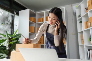 Genç bir girişimci telefonda konuşurken, paketleri parlak bir ofis alanında hazırlıyor, çoklu görev ve iş zekasını vurguluyor.