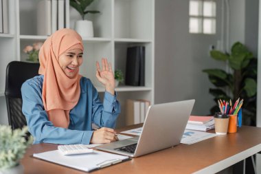Çağdaş bir ofis ortamında video çağrısına katılan tesettür dalgaları takan neşeli Müslüman bir kadın....
