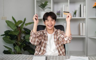 Neşeli bir genç elinde bir kredi kartı tutuyor ve başarılı online alışverişini kutluyor. Heyecan ve başarıyı ifade ediyor..