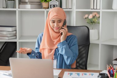 Başörtüsü takan Müslüman bir kadın, masasında çalışırken yanında mali raporlar ve dizüstü bilgisayarla telefon görüşmesi yapıyor..