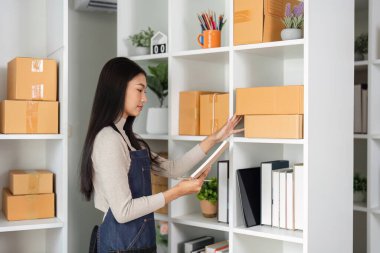 Modern bir ofiste raftaki paketleri yönetirken elinde tabletle envanter düzenleyen bir iş kadını..