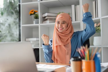 Modern bir ofiste masasında profesyonel bir başarı elde ettikten sonra tesettürlü bir Müslüman kadın yumruklarıyla sevinç ve sevinç ifade ediyor..