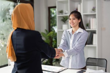 Çağdaş bir ofiste el sıkışırken iki farklı iş kadını gülümsüyor, işbirliği ve profesyonel ilişkilerinde güçlü bir ortaklık vurguluyor.