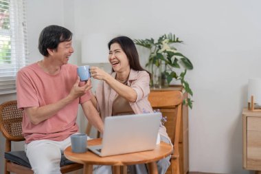 Mutlu yaşlı bir çift, rahat evlerinde çevrimiçi aktivitelerin keyfini çıkarırken kahve fincanlarıyla neşeli bir kadeh kaldırır..