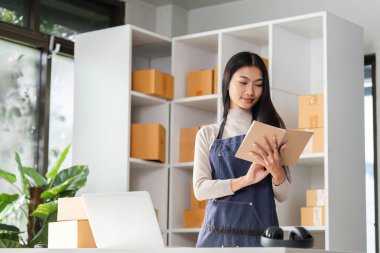 Önlüklü genç bir kadın, kutularla dolu, parlak ve düzenli bir çalışma alanında mal sevkiyatını yönetmek ve takip etmek için tablet kullanıyor..