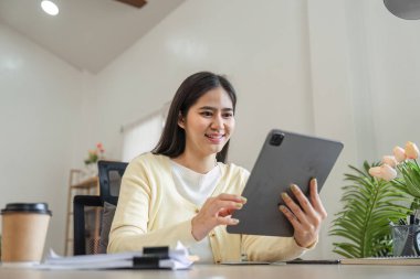 Gülümseyen genç bir kadın, teknoloji ve verimliliğin bir karışımını yansıtan, rahat bir ev ofisinde tabletiyle etkileşime girer..