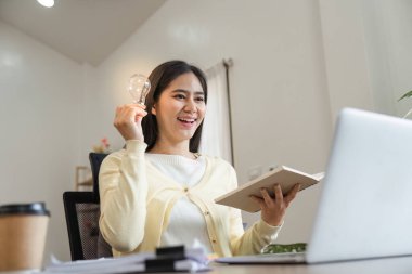 Neşeli genç bir kadın, dizüstü bilgisayarıyla uğraşırken bir elinde ampul tutuyor. Çalışma alanındaki yaratıcılığı ve ilhamı simgeliyor..