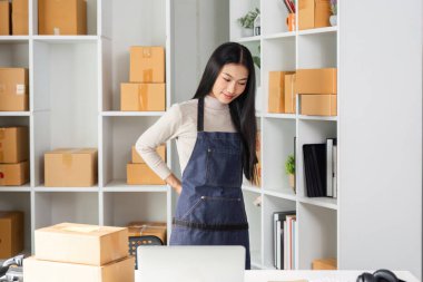 Önlüklü genç bir kadın envanter düzenliyor ve kutularla ve raflarla dolu modern bir çalışma alanında nakliyat yönetiyor..