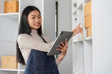 Envanteri yönetmek için tablet kullanan ve modern ofis çalışma alanındaki raflardaki paketlere işaret eden bir e-ticaret işkadını..