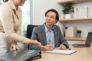 Bir iş adamı bir meslektaşıyla görüşürken belgeleri inceler ve imzalar, işbirliğini ve ayrıntılara olan ilgiyi vurgular.