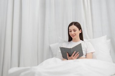 Yumuşak ışıklandırma ve rahat yatak örtüsüyle çevrili, rahat bir yatak odasında dinlenen bir kadının huzurlu bir görüntüsü, huzurlu bir atmosfer yaratıyor..