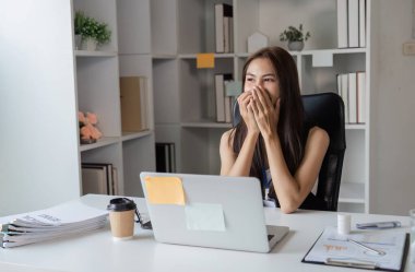 Modern ofis masasında neşeli bir iş kadını. Dizüstü bilgisayarı üzerinde çalışırken gülümsüyor ve heyecanını dile getiriyor. Yakınlarda bir kahve fincanı ve belgeler var..