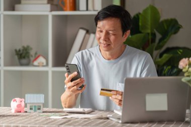 Gülümseyen son sınıf öğrencisi online alışveriş için akıllı telefon kullanıyor. Ev ortamında dizüstü bilgisayar ve dekoratif eşyalarla otururken elinde kredi kartı tutuyor..