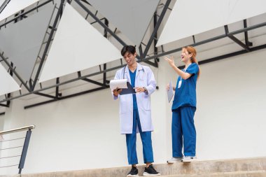 Önlüklü ve laboratuvar önlüklü genç bir doktor çağdaş bir binanın dışındaki hasta vakalarını inceleyerek sağlık ve modern mimarinin bir karışımını sergiliyor..