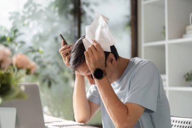 Günlük giysiler içinde başını mendille tutan genç bir adam, evde dizüstü bilgisayarlı bir masada otururken mali kararların verdiği stresi dile getiriyor..