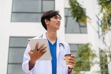 Binanın dışında duran genç bir doktor elinde kahve fincanı tutarken tabletteki hasta vakalarını kontrol ediyor..