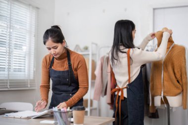 Kendini işine adamış iki kadın girişimci online gömlek işlerini yürütüyor, biri elbise asarken diğeri şık bir butik çalışma alanında notlar alıyor..