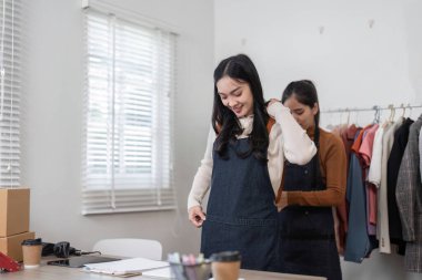 Parlak ve düzenli bir çalışma alanında online gömlek satışları için hazırlanan iki neşeli bayan girişimci şık önlüklere sığar..