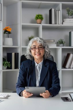 Kitaplar ve bitkilerle çevrili, elinde tabletle modern bir ofiste oturan, kendine güveni tam yaşlı bir iş kadını..