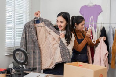 İki dinamik bayan girişimci, çağdaş online gömlek butik çalışma alanlarında giysi eşyaları düzenlerken şık bir palto sergiliyorlar..