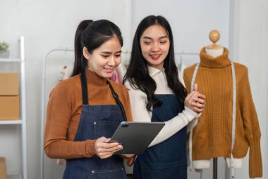İki yetenekli kadın moda tasarımcısı bir tablete bakıyor, son tasarımlarını tartışıyorlar butikte özel dikim bir kıyafet sergisinin yanında dururken.