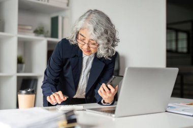 Yaşlı bir iş kadını, elinde akıllı telefon tutarken hesap makinesi kullanarak, modern ofis alanında verimlilik göstererek birden fazla iş yapmakla meşgul..