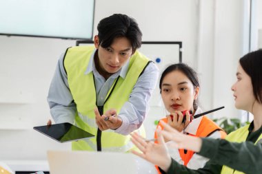 Güvenlik yeleği giymiş bir inşaat yöneticisi, bir tabletle ekiple temas kuruyor, bir işbirliği oturumu sırasında proje planları ve yönergeler hakkında ayrıntılı bir tartışma yürütüyor..