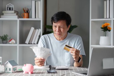 Mali durumunu değerlendiren, faturaları ve kredi kartını elinde tutan endişeli bir adam. Evrak işleriyle dolu evinde endişelerini dile getiriyor..