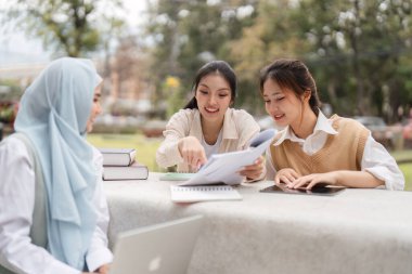 Bir grup farklı üniversite öğrencisi açık havada birlikte çalışıyor, akademik materyalleri tartışıyor ve bir çalışma sırasında görüşlerini paylaşıyorlar..