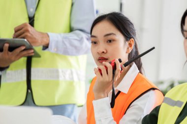Turuncu yelek giymiş, takım üyeleriyle koordine etmek için telsiz kullanan, proje infazı sırasında etkili bir iletişim sağlayan odaklanmış bir inşaat denetçisi..