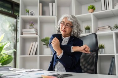 İş elbisesi giymiş yaşlı bir kadın masasında çalışırken nazikçe omzunu uzatıyor ve çağdaş ofis ortamında kendine bakıcılık yapıyor..