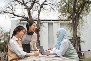 Ağaçlar ve akademik materyallerle çevrili, dışarıda çalışırken gülen ve işbirliği yapan çeşitli bir üniversite öğrencisi grubu.