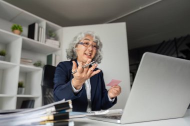 Hevesli, yaşlı bir iş kadını, video görüşmesi sırasında elinde bir notla, zarif bir çalışma alanında düşüncelerini tutkuyla paylaşıyor..