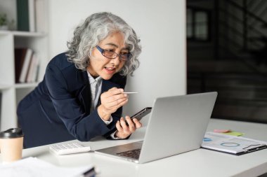 Tecrübeli bir iş kadını bilgisayarındaki verileri incelerken aktif olarak akıllı telefonunu tutar ve şık bir ofis ortamında dinamik bir etkileşim gösterir..