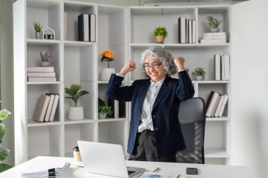 Hevesli yaşlı bir iş kadını, modern masasında otururken kollarını zaferle kaldırarak profesyonel bir başarıyı kutluyor..