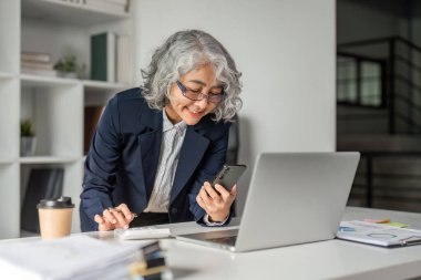 Yaşlı bir iş kadını, çağdaş bir ofis ortamında dokümanlar ve dizüstü bilgisayarla çevrili akıllı telefonunu kontrol ederken bir anlık pozitifliğin tadını çıkarıyor..