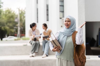 Kitapları tutarken gülümseyen ve el sallayan çeşitli bir kız öğrenci, sınıf arkadaşlarıyla birlikte kampüsün arka planında bir çalışma oturumundaydılar..