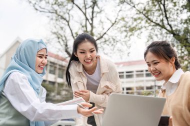 Üç çok kültürlü kız öğrenci, açık havada canlı bir akademik ortamda fikirlerini paylaşırken bir dizüstü bilgisayarda gülümsüyor ve işbirliği yapıyorlar..