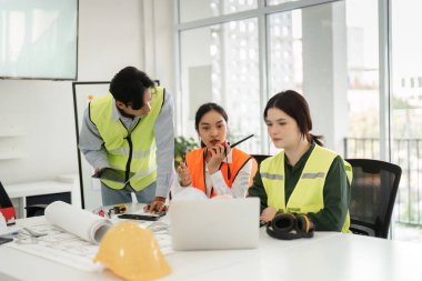 Şık, çağdaş bir ofiste işbirliği yaparken dizüstü bilgisayardaki planları inceleyen bir mimar, mühendis ve tasarımcıdan oluşan dinamik bir inşaat ekibi..