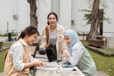 Bir grup çok kültürlü kadın, bir üniversite ortamında açık havada tartışırken dizüstü bilgisayar ve defterlerle bir proje üzerinde işbirliği yapıyorlar..