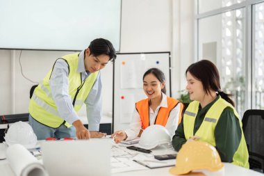Güvenlik yeleği ve kask giyen üç inşaat işçisi, çağdaş bir ofis planlarını tartışmak ve analiz etmek için ortak bir toplantıya giriştiler..